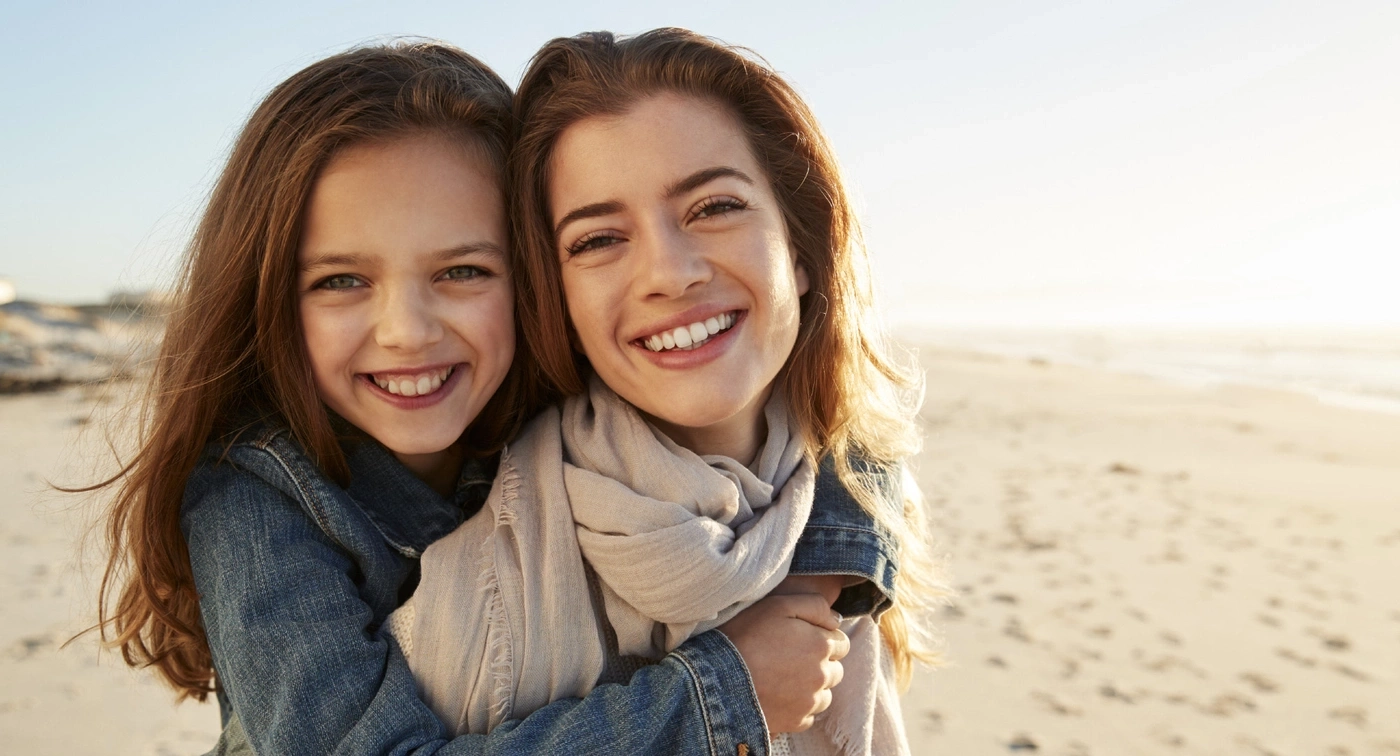 Zwei lächelnde Frauen umarmen sich am Strand, glücklich und sonnig.